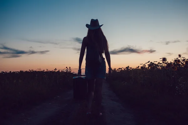 Mädchen mit Cowboyhut steht mit einem Koffer auf der Straße im Sonnenblumenfeld. Warten auf Hilfe. Sonnenuntergang. — Stockfoto