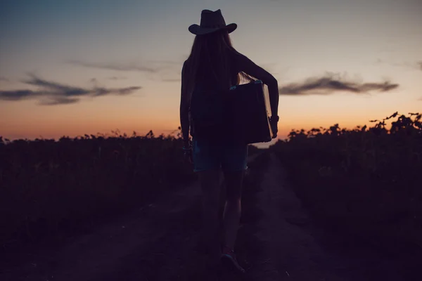 Mädchen mit Cowboyhut steht mit einem Koffer auf der Straße im Sonnenblumenfeld. Warten auf Hilfe. Sonnenuntergang. — Stockfoto