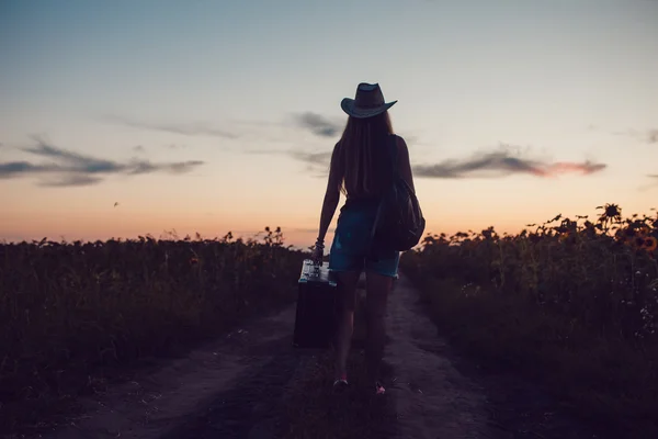 Fille dans un chapeau de cow-boy debout avec une valise sur la route dans le champ de tournesol. J'attends de l'aide. Coucher de soleil . — Photo