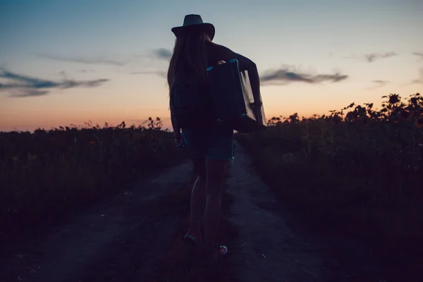 Meisje in een staande cowboy hoed met een koffer op de weg in de zonnebloem veld. Wachten op hulp. Zonsondergang. — Stockfoto