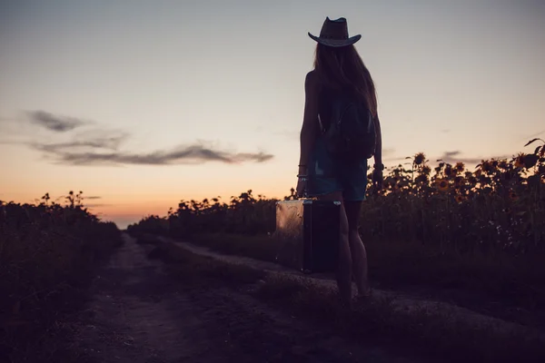 Flicka i en cowboyhatt stående med en resväska på vägen i fältet solros. Väntar på hjälp. Solnedgång. — Stockfoto