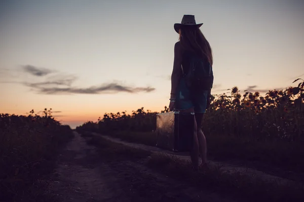 Mädchen mit Cowboyhut steht mit einem Koffer auf der Straße im Sonnenblumenfeld. Warten auf Hilfe. Sonnenuntergang. — Stockfoto