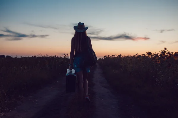 Mädchen mit Cowboyhut steht mit einem Koffer auf der Straße im Sonnenblumenfeld. Warten auf Hilfe. Sonnenuntergang. — Stockfoto
