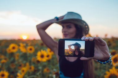 Kovboy şapkalı bir bavulun ayçiçeği bir alan içinde oturan kız. Günbatımı.