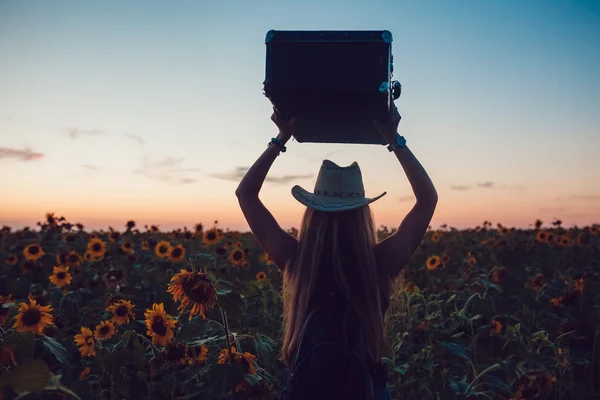 Meisje in een staande cowboy hoed met een koffer op de weg in de zonnebloem veld. Wachten op hulp. Zonsondergang. — Stockfoto