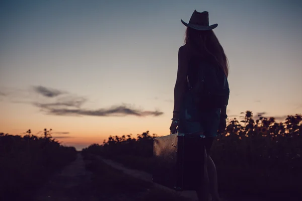 Meisje in een staande cowboy hoed met een koffer op de weg in de zonnebloem veld. Wachten op hulp. Zonsondergang. — Stockfoto