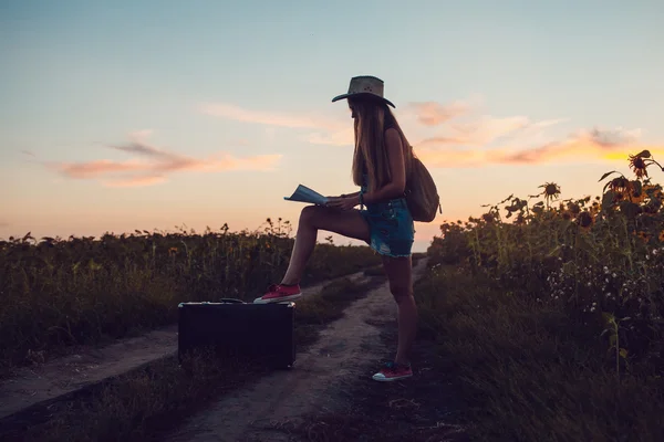 Meisje in cowboy hoed zittend op een koffer op een zonnebloem gebied. Zonsondergang. — Stockfoto