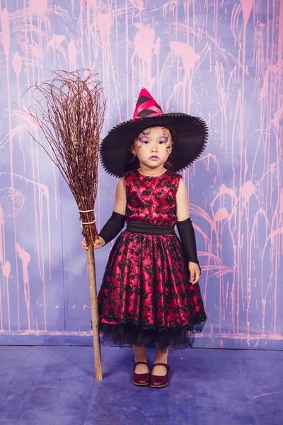 Menina vestida de bruxa com vassoura em pé no fundo da parede roxa — Fotografia de Stock