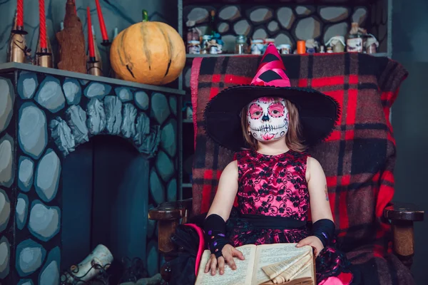少女の魔女の衣装は、ハロウィーンの飾りで彼の手の本と椅子に座って彼女の顔にメイクアップ — ストック写真