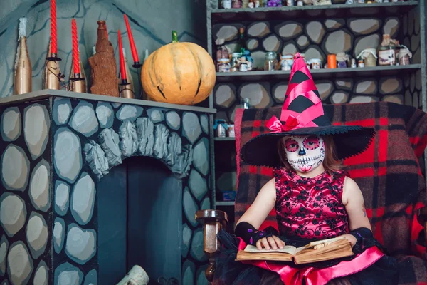 Девушка в костюме ведьмы и макияж на ее лице сидя в кресле с книгой в руках в Хэллоуин украшения — стоковое фото