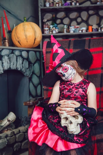 Menina em um traje de bruxa sentado na cadeira com o crânio em suas mãos em decorações halloween — Fotografia de Stock