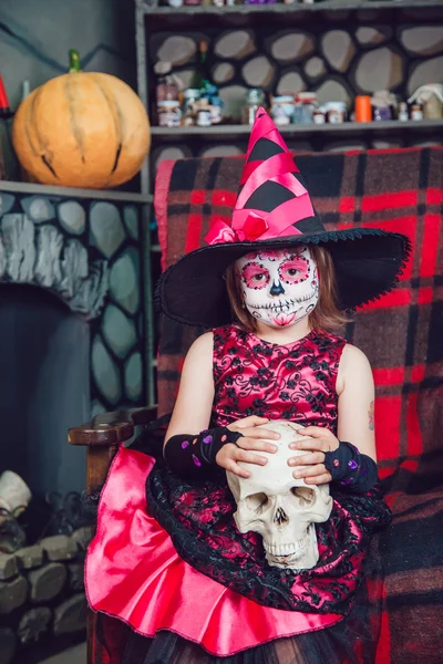 Mädchen im Hexenkostüm auf Stuhl sitzend mit Totenkopf in den Händen in Halloween-Dekoration — Stockfoto