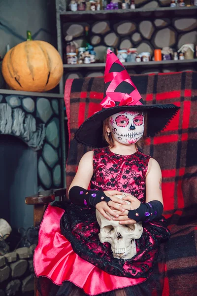 Menina em um traje de bruxa sentado na cadeira com o crânio em suas mãos em decorações halloween — Fotografia de Stock