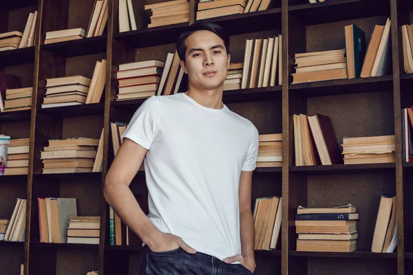 Asiatisk man i vit T-shirt står mot bakgrund av bokhyllor. Uppställning. — Stockfoto