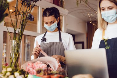 Koruyucu maskeli çiçekçi işçileri bir buket süsleyip dizüstü bilgisayar kullanıyorlar..
