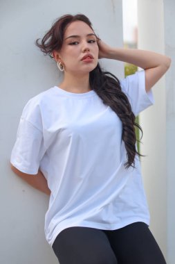 Beyaz tişörtlü ve siyah taytlı şık genç bir kadın yemyeşil çimlerin arasında rahatça poz veriyor.