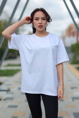 Kendine güvenen genç bir kadın şık, büyük boy beyaz bir tişörtle poz veriyor. Zarif bir şekilde şık taytlarla eşleştirilmiş.