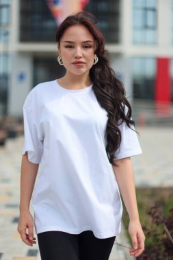 Genç bir kadın şık beyaz bir tişörtle canlı bir modern mimari zemine karşı güvenle poz veriyor.