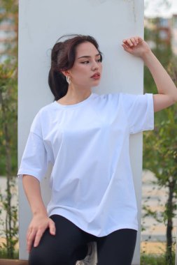 Beyaz tişörtlü ve siyah taytlı şık genç bir kadın yemyeşil çimlerin arasında rahatça poz veriyor.