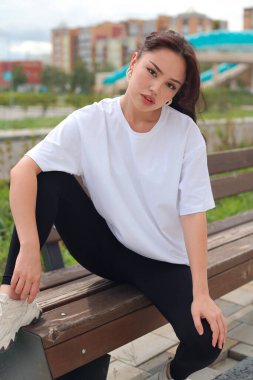 Şık bir genç kadın şehir ortamında bir bankta oturuyor, gündelik moda ve modern bir hava sergiliyor..