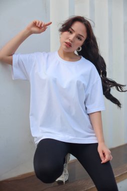 Beyaz tişörtlü ve siyah taytlı şık genç bir kadın yemyeşil çimlerin arasında rahatça poz veriyor.