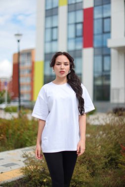 Genç bir kadın şık beyaz bir tişörtle canlı bir modern mimari zemine karşı güvenle poz veriyor.