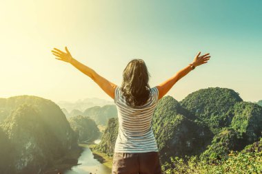 Uçurumun kenarında duran ve ellerini yüksek kayalık dağlara doğru kaldıran çekici bir kadın Tam Coc, Ninh Binh Eyaleti, Vietnam 'ın güzel manzarasına hayran kalıyor..