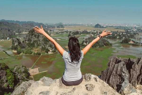 Uçurumun kenarında duran ve ellerini yüksek kayalık dağlara doğru kaldıran çekici bir kadın Tam Coc, Ninh Binh Eyaleti, Vietnam 'ın güzel manzarasına hayran kalıyor..