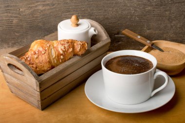 Kahveli beyaz fincan, ahşap bir kutuda kruvasan ve süt kabı..