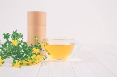 Bir fincan taze bitkisel antioksidan çayı, Hypericum çiçekleri ve beyaz masanın üzerinde boş kağıt ambalaj tüpü. Alternatif tıp, şifalı bitki ve sağlık konsepti. Sıfır atık çay ambalajı, model resim