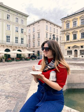 Tarihi şehir merkezinde fıskiyenin yanında oturan ve sabah kahvesinin keyfini çıkaran genç ve güzel bir kadının yakın çekimi. Yerel turizm ve seyahat konsepti. Seçici odak, biçimlendirme resmi