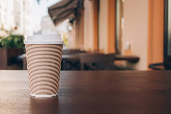 Mockup image of paper cup of coffee take-away, standing on wooden table with blurred cafe behind - as background with copyspace. Coffee to go in recyclable paper cup