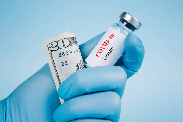 Doctor or scientist holding 20 dollars bill and vial dose of COVID-19 vaccine against blue background with copy space - global vaccination and money laundering concept, vaccine cost. Selective focus