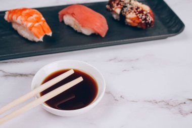 Soya soslu yemek çubukları ve ton balıklı suşi nigiri, karides ve füme yılan balığı beyaz mermer masa arkasında siyah tabakta. Suşi bar menüsü, yemek teslimatı restoran yemeği konsepti