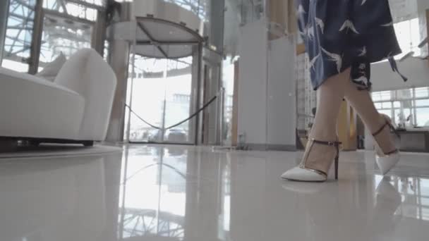 Ένα κορίτσι με ψηλά τακούνια περπατάει στο διάδρομο ενός επιχειρηματικού κέντρου ή ξενοδοχείου. Λευκά παπούτσια και μπλε φόρεμα. — Αρχείο Βίντεο