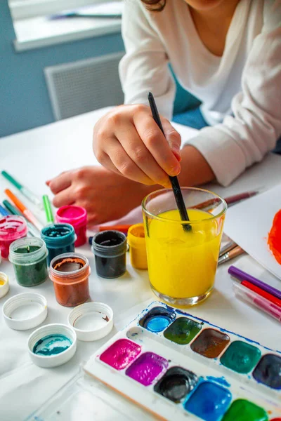 Das Kind senkt seine Hand ins Deltawasser und wäscht die Gouache ab. Hausaufgabenbetreuung. Zeichentraining. Unabhängigkeit der Kinder von der Kunst. — Stockfoto