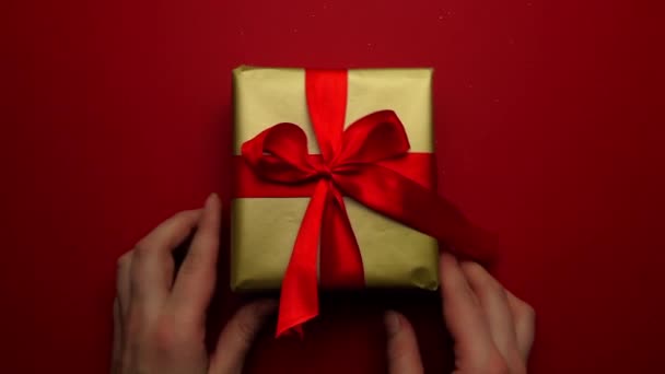 Праздничный подарок в золотой упаковке и красный бант на красном фоне. Крупным планом девушки кладут подарок на стол и выпрямляют бант на подарке. Приятный подарок и сюрприз для близких. — стоковое видео