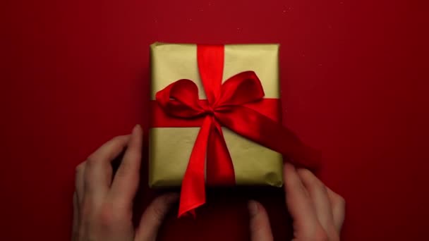 Regalo festivo in confezione d'oro e fiocco rosso su fondo rosso. Primo piano, le mani delle ragazze mettono un regalo sul tavolo e raddrizzano l'arco sul regalo. Un piacevole regalo e sorpresa per i propri cari. — Video Stock