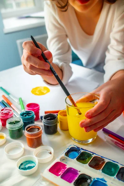 Das Kind senkt seine Hand ins Deltawasser und wäscht die Gouache ab. Hausaufgabenbetreuung. Zeichentraining. Unabhängigkeit der Kinder von der Kunst. — Stockfoto