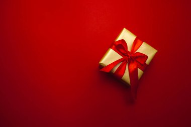 Tatil hediyesi. Kırmızı bir arka planda kırmızı bir fiyonkla paketlenmiş altın bir hediye kutusu. Mutlu Noeller, mutlu yıllar ve sevgililer günü selamları. Kartpostal