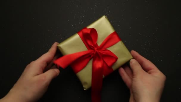 Εορταστικό δώρο σε χρυσή συσκευασία και ένα κόκκινο φιόγκο σε μαύρο φόντο. Κοντινό πλάνο, τα χέρια των κοριτσιών βάζουν ένα δώρο σε μαύρο φόντο και ισιώνουν το τόξο στο δώρο. Ένα ευχάριστο δώρο και έκπληξη για — Αρχείο Βίντεο