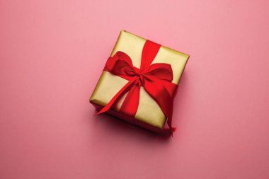 Tatil hediyesi. Pembe arka planda kırmızı kurdeleyle paketlenmiş altın bir hediye kutusu. Mutlu Noeller Sevgililer Günü. Sevgi dolu kartpostal.