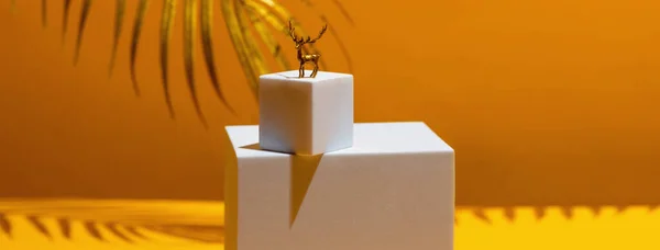 En lille figur af en gylden hjort et symbol på det nye år på to hvide firkanter, i stil med monumentalisme, på en gul baggrund, bag bladene fra et palmetræ - Stock-foto