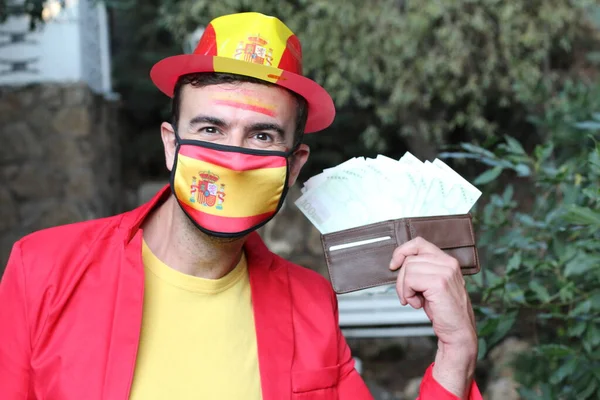 portrait of handsome young man in clothes colored in Spain flag and medical mask holding wallet full of money on nature