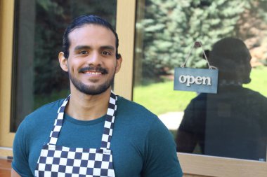 Kapının önünde açık hesap ile damalı önlüklü yakışıklı Latin küçük işletme sahibinin portresi.