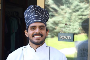 Yakışıklı Latin aşçının portresi. Üniformalı ve şapkalı. Restoran kapısının önünde.