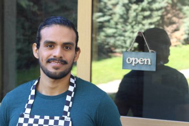 Kapının önünde açık hesap ile damalı önlüklü yakışıklı Latin küçük işletme sahibinin portresi.