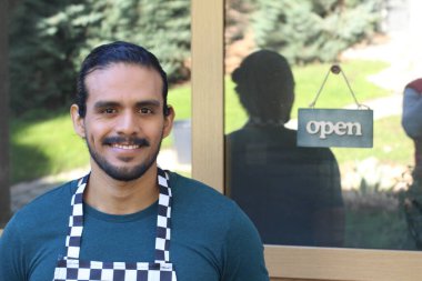 Kapının önünde açık hesap ile damalı önlüklü yakışıklı Latin küçük işletme sahibinin portresi.