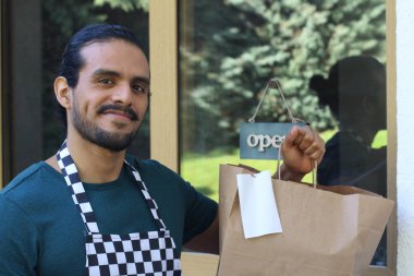 Kapının önünde kağıt torba ve açık hesap ile yakışıklı Latin üniformalı küçük işletme sahibi portresi