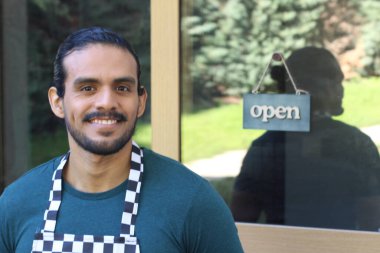 Kapının önünde açık hesap ile damalı önlüklü yakışıklı Latin küçük işletme sahibinin portresi.